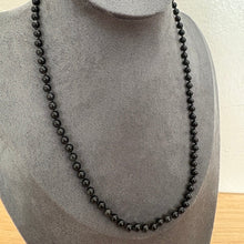 Load image into Gallery viewer, A long black beaded necklace displayed on a grey fabric jewellery bust against a light wall and wood surface.
