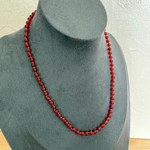 Load image into Gallery viewer, A single strand red beaded necklace displayed on a dark grey fabric jewellery bust against a light textured wall.
