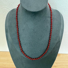 Load image into Gallery viewer, A long strand of dark red polished gemstone beads, a beaded necklace, displayed on a dark grey velvet jewellery bust against a white wall and light wood surface.
