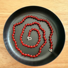 Load image into Gallery viewer, A red beaded necklace with a silver clasp, arranged in a spiral on a black circular tray over a light wood background.
