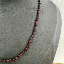 Load image into Gallery viewer, A dark red beaded necklace displayed on a grey fabric jewellery bust against a light wall.
