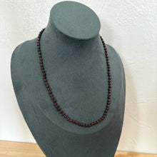 Load image into Gallery viewer, A dark red beaded necklace displayed on a dark grey velvet jewellery bust against a white textured wall.
