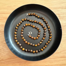Load image into Gallery viewer, A beaded necklace made of round brown and golden-brown beads with a silver bolt clasp, arranged in a spiral on a black tray over a light wood surface.
