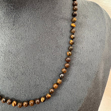 Load image into Gallery viewer, A beaded necklace made of polished brown and tan tiger's eye beads, displayed on a dark grey fabric jewellery bust.
