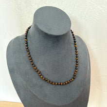 Load image into Gallery viewer, A beaded necklace made of small brown and tan round beads, displayed on a dark grey velvet jewellery bust against a light textured wall.
