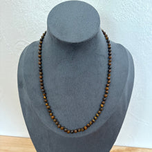 Load image into Gallery viewer, A beaded necklace made of small brown and tan tiger's eye beads, displayed on a dark grey velvet jewellery bust against a white textured wall.
